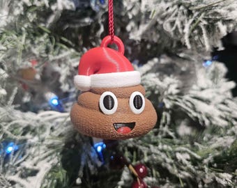 May include: A brown poop emoji Christmas ornament wearing a red Santa hat with white trim. The ornament has large, round eyes and a smiling mouth. It hangs from a red string against a snowy Christmas tree background.