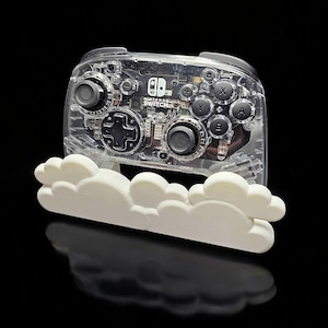 May include: A clear plastic Nintendo Switch controller with black buttons and a white cloud-shaped stand. The controller has the Nintendo Switch logo on the front.