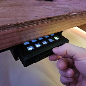 May include: A black streaming control panel with buttons for play, pause, mute, and other functions. The panel has a white "STREAM" label and is mounted under a wooden desk.