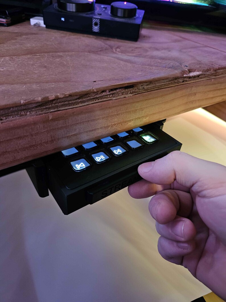 May include: A black streaming control panel with buttons for play, pause, mute, and other functions. The panel has a white "STREAM" label and is mounted under a wooden desk.