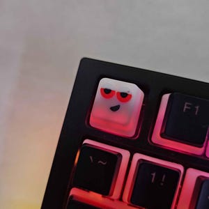 May include: A white keyboard keycap with a red and white cartoon face. The face has two red eyes, a black mouth, and a white eyebrow.