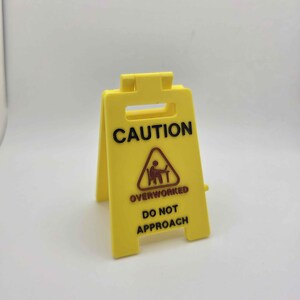 May include: A yellow plastic caution sign with the word "CAUTION" in black capital letters. Below, a triangle with a person at a desk is above the words "OVERWORKED DO NOT APPROACH". The sign is freestanding.