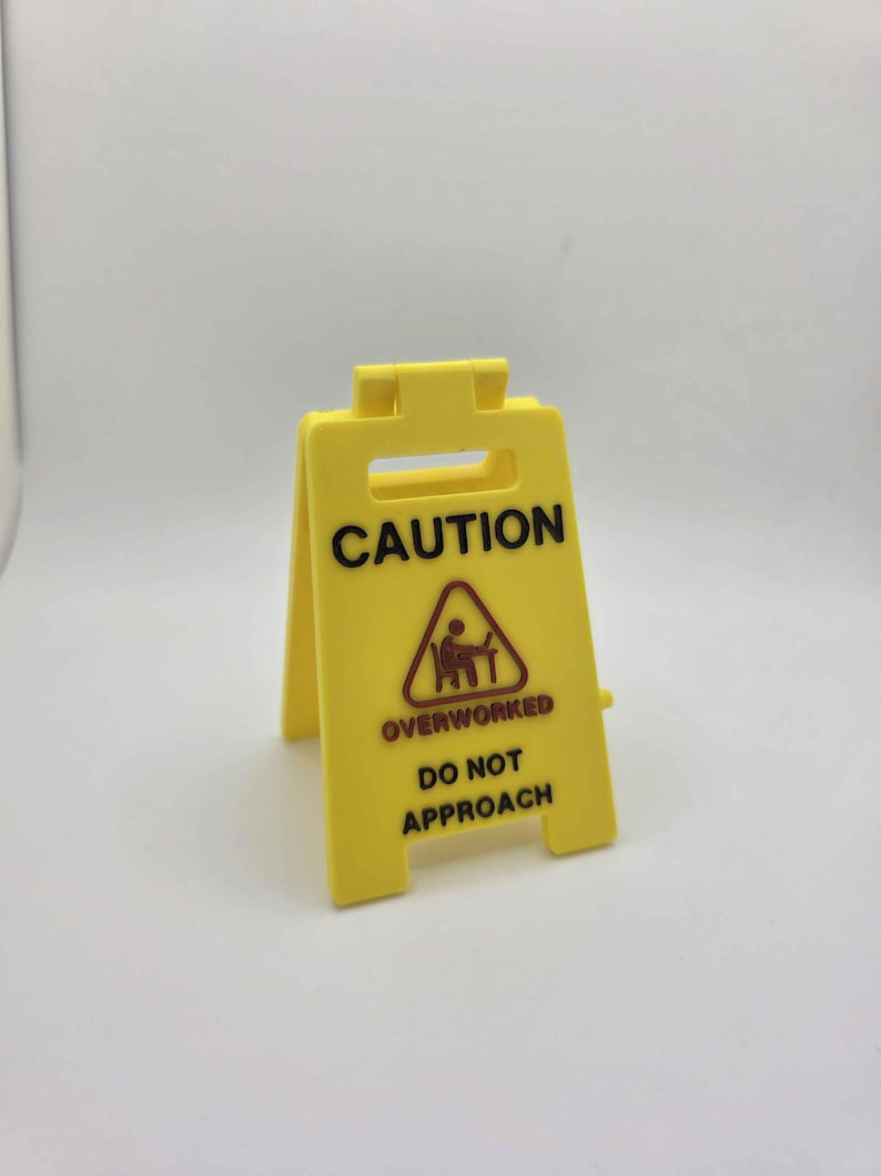 May include: A yellow plastic caution sign with the word "CAUTION" in black capital letters. Below, a triangle with a person at a desk is above the words "OVERWORKED DO NOT APPROACH". The sign is freestanding.