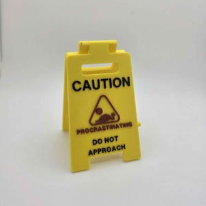 May include: A yellow plastic caution sign with black text that reads "CAUTION PROCRATINATING DO NOT APPROACH". The sign has a brown and red graphic of a person in a triangle.