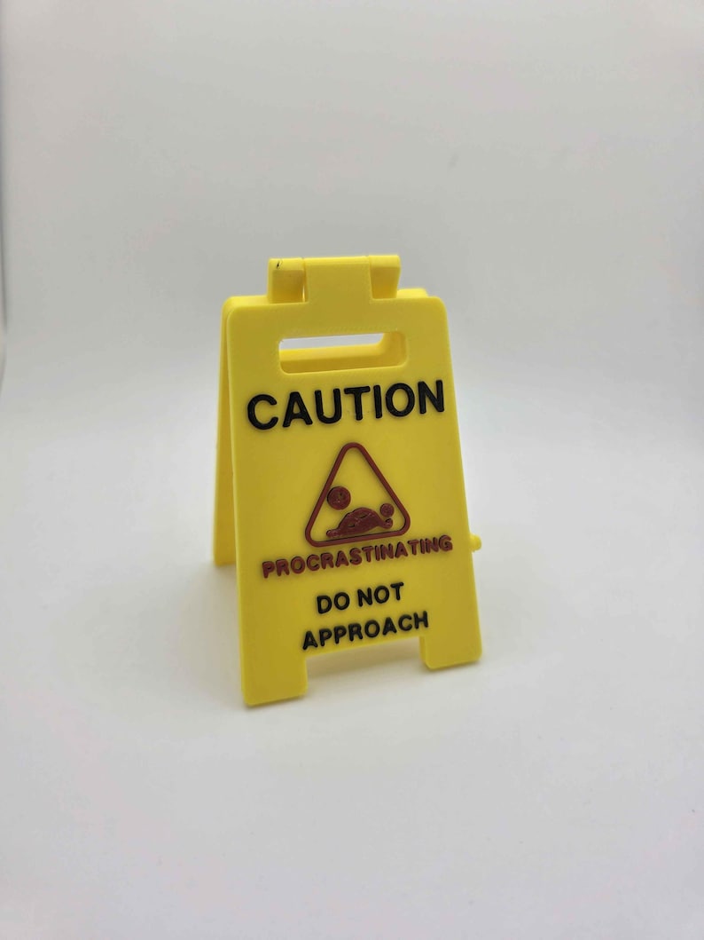 May include: A yellow plastic caution sign with black text that reads "CAUTION PROCRATINATING DO NOT APPROACH". The sign has a brown and red graphic of a person in a triangle.
