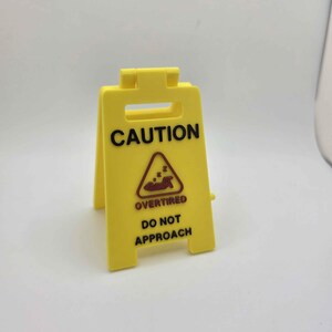May include: A yellow plastic caution sign with the words "CAUTION" at the top. Below is a graphic of a person sleeping and the words "OVERTIRED DO NOT APPROACH". The sign is a-frame style and stands upright.