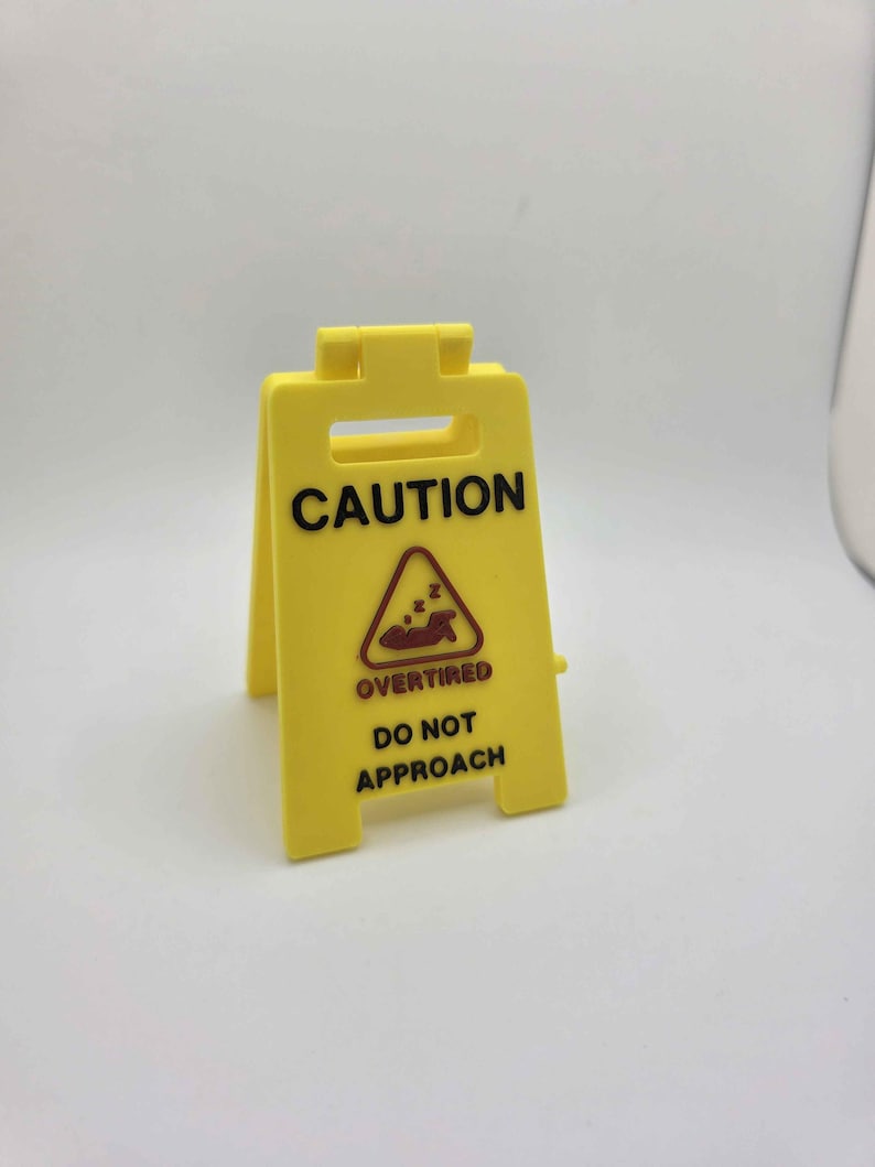 May include: A yellow plastic caution sign with the words "CAUTION" at the top. Below is a graphic of a person sleeping and the words "OVERTIRED DO NOT APPROACH". The sign is a-frame style and stands upright.
