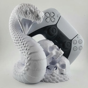May include: A white 3D-printed gaming controller stand featuring a snake coiled around two skulls. A white and black gaming controller rests on the stand. The stand is a decorative accessory for gamers.