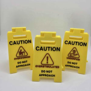 May include: Three yellow caution signs with black text and symbols. Each sign reads "CAUTION" at the top. The signs warn of being "OVERWORKED", "OVERSTIMULATED", and "UNDERPAID", with "DO NOT APPROACH" below each warning.