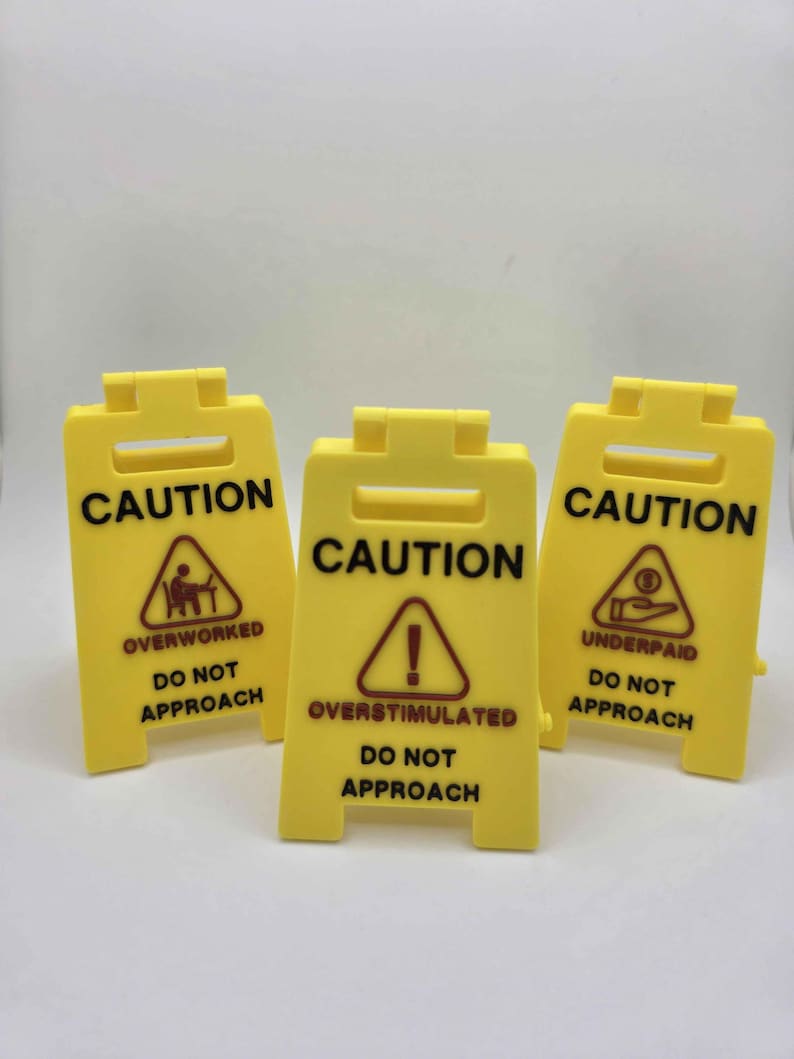 May include: Three yellow caution signs with black text and symbols. Each sign reads "CAUTION" at the top. The signs warn of being "OVERWORKED", "OVERSTIMULATED", and "UNDERPAID", with "DO NOT APPROACH" below each warning.
