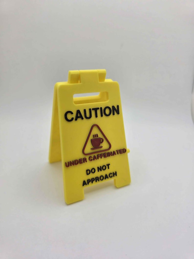 May include: A yellow plastic caution sign with black text that reads "CAUTION UNDER CAFFEINATED DO NOT APPROACH." The sign features a graphic of a coffee cup with steam. The sign is folded in a triangular shape and stands upright.