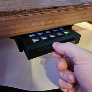 May include: A black keyboard with white keys and a green button. The keyboard is mounted under a wooden desk and a hand is reaching for the green button.