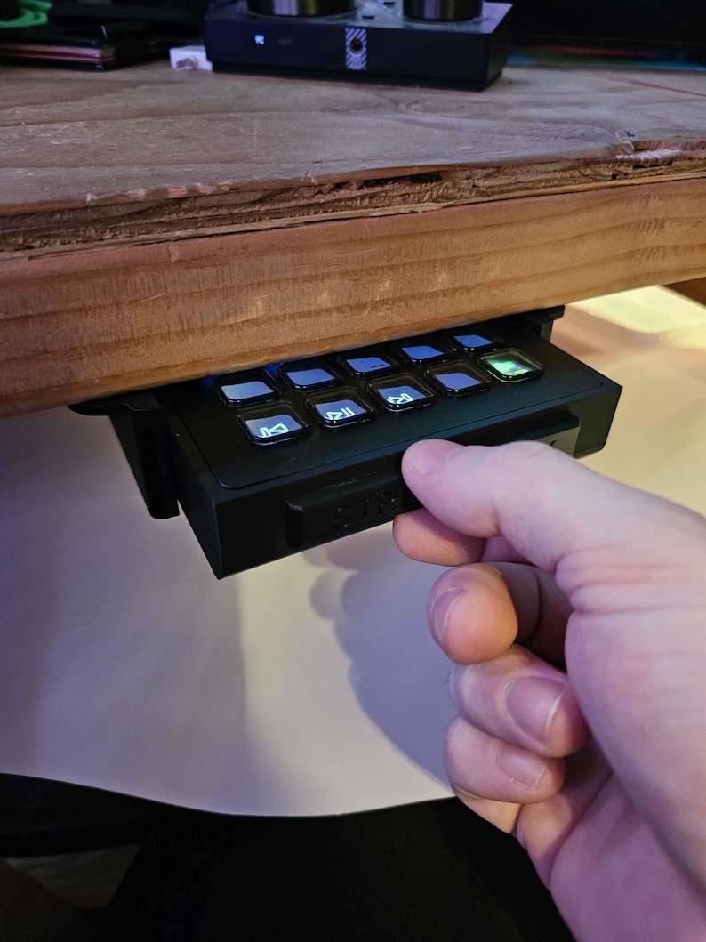 May include: A black keyboard with white keys and a green button. The keyboard is mounted under a wooden desk and a hand is reaching for the green button.