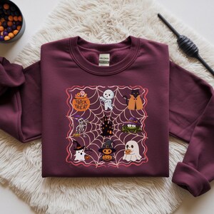 以下が含まれることがあります： ハロウィーンをテーマにしたグラフィックが特徴のマルーンのスウェットシャツ。グラフィックには、幽霊、猫、カボチャなどのさまざまな漫画のキャラクターが描かれたクモの巣のデザインが含まれています。キャンディーのボウルが左上にあります。