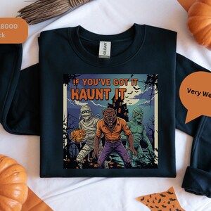 Könnte beinhalten: Schwarzes Sweatshirt mit einem Halloween-Motiv. Das Motiv zeigt eine Mumie, einen Werwolf und eine Kreatur aus der schwarzen Lagune mit dem Text "If You've Got It Haunt It". Das Sweatshirt hat einen Rundhalsausschnitt und lange Ärmel.