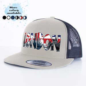 Puede incluir: Gorra snapback gris claro con parte trasera de malla azul marino. La palabra "LONDON" está impresa en la parte delantera con una fuente estilizada con un diseño de la Union Jack y un fondo de paisaje urbano. El texto "Más colores disponibles" está arriba.