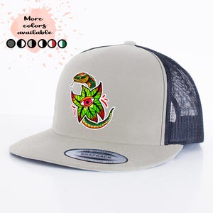 May include: A light gray and navy blue trucker hat with a colorful graphic of a snake and an eye on a green leaf. The hat has a mesh back and a curved brim. The text "More colors available" is above the hat.