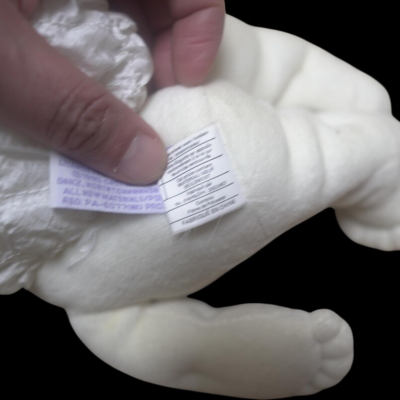 Puede incluir: Un juguete de peluche blanco roto, parcialmente visible, sostenido por una mano. La textura del juguete parece suave. Una etiqueta con texto est&aacute; adherida al juguete. El fondo es negro.