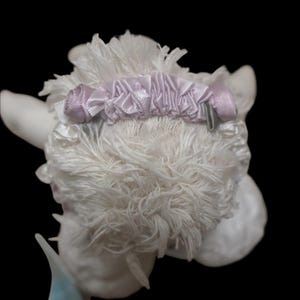 Puede incluir: Figura decorativa de cordero blanco y esponjoso con una cinta y un lazo de sat&eacute;n morado claro alrededor de la cabeza. El cordero tiene un aspecto texturizado, similar a la lana, sobre un fondo negro.