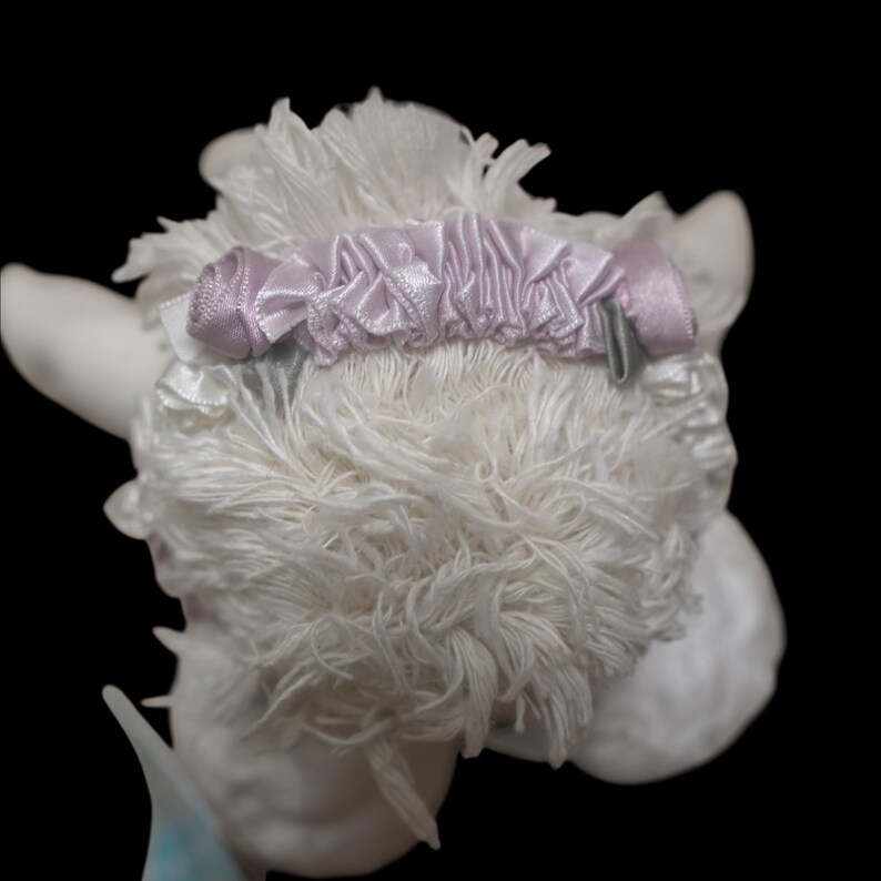 Puede incluir: Figura decorativa de cordero blanco y esponjoso con una cinta y un lazo de sat&eacute;n morado claro alrededor de la cabeza. El cordero tiene un aspecto texturizado, similar a la lana, sobre un fondo negro.