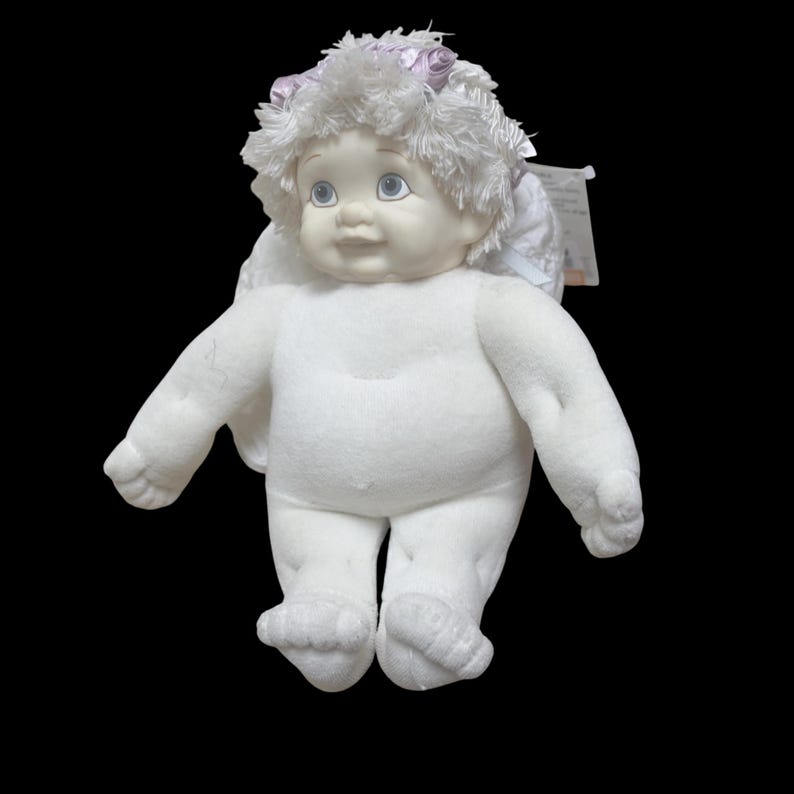 Puede incluir: Una mu&ntilde;eca de &aacute;ngel de felpa blanca con una cara pintada, ojos azules y un halo de pelusa blanca y detalles en rosa. La mu&ntilde;eca tiene alas blancas adjuntas y est&aacute; sobre un fondo negro.