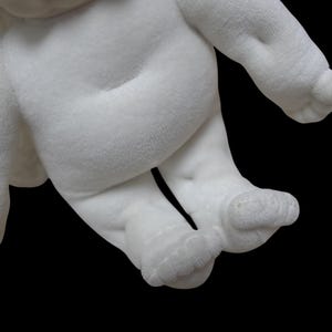 Puede incluir: Primer plano de un juguete de peluche blanco, que muestra el cuerpo, las piernas y los pies. La tela suave y texturizada es visible, con detalles como el ombligo y la forma de los pies. El fondo es negro liso.
