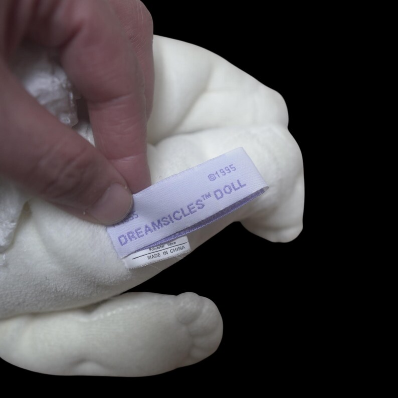 Puede incluir: Una mu&ntilde;eca de peluche blanca con una etiqueta que dice "DREAMSICLES DOLL" y "&copy;1995". La etiqueta tambi&eacute;n indica que la mu&ntilde;eca est&aacute; hecha de fibra de poli&eacute;ster y fabricada en China. La mu&ntilde;eca se sostiene sobre un fondo negro.