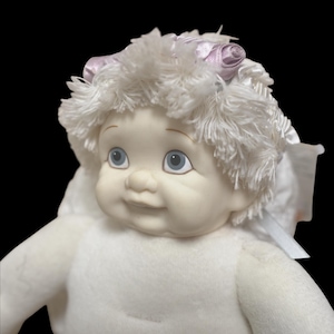 Puede incluir: Una mu&ntilde;eca de peluche con una cara sonriente, ojos azules y cabello blanco. La mu&ntilde;eca tiene un cuerpo blanco y est&aacute; adornada con un lazo rosa. La mu&ntilde;eca est&aacute; sobre un fondo negro.