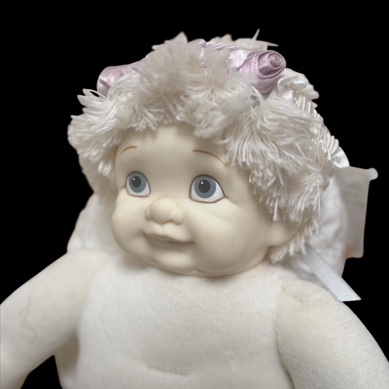 Puede incluir: Una mu&ntilde;eca de peluche con una cara sonriente, ojos azules y cabello blanco. La mu&ntilde;eca tiene un cuerpo blanco y est&aacute; adornada con un lazo rosa. La mu&ntilde;eca est&aacute; sobre un fondo negro.