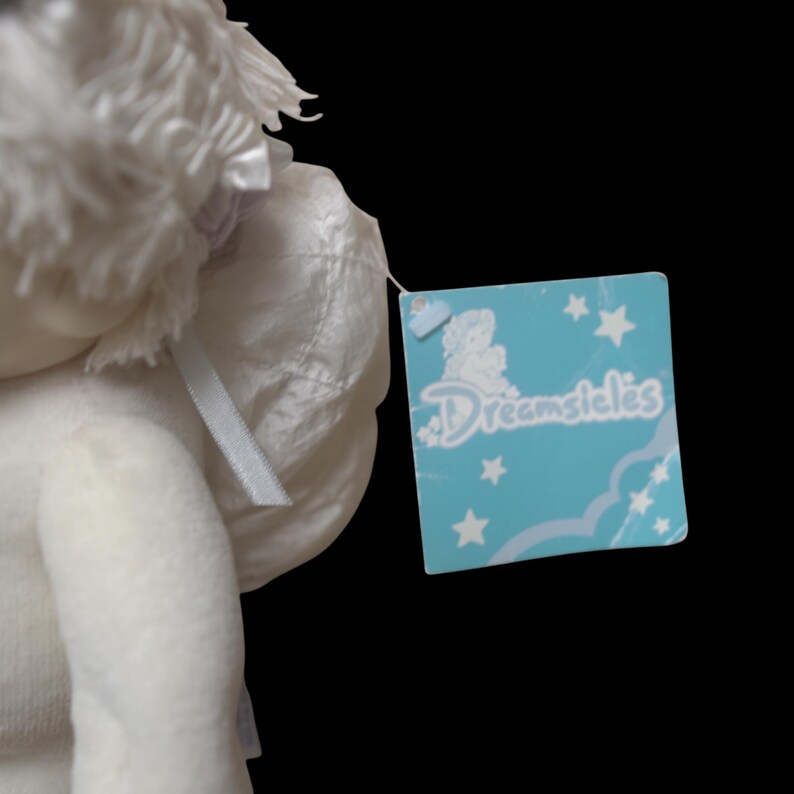 Puede incluir: Una mu&ntilde;eca de &aacute;ngel de peluche blanco con una etiqueta que dice "Dreamsicles". La mu&ntilde;eca tiene alas blancas y una cinta. La etiqueta es azul claro con estrellas blancas y un dise&ntilde;o de nube.