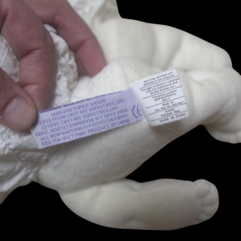 Puede incluir: Primer plano de un juguete de peluche blanco con una etiqueta adjunta. La etiqueta dice "MANUFACTURED UNDER LICENSE FROM CAST ART INDUSTRIES INC." e incluye detalles sobre los materiales y el origen. El juguete est&aacute; hecho de fibras de poli&eacute;ster.
