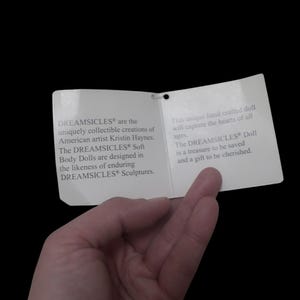 Puede incluir: Una etiqueta de papel blanco con texto sobre las mu&ntilde;ecas DREAMSICLES&reg;, sostenida por una mano sobre un fondo negro. La etiqueta describe las mu&ntilde;ecas como creaciones coleccionables &uacute;nicas de la artista estadounidense Kristin Haynes.
