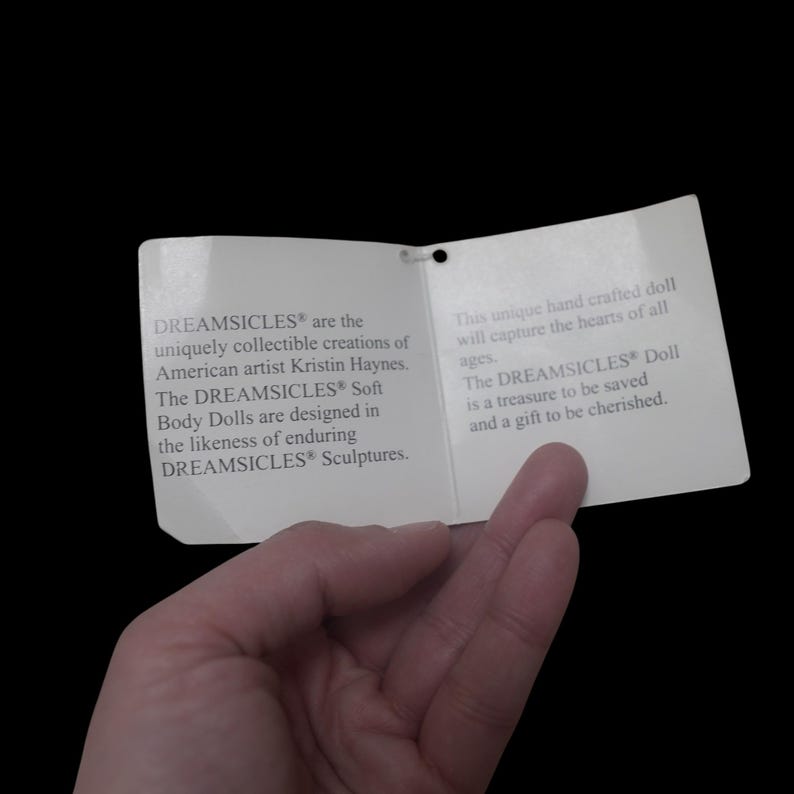 Puede incluir: Una etiqueta de papel blanco con texto sobre las mu&ntilde;ecas DREAMSICLES&reg;, sostenida por una mano sobre un fondo negro. La etiqueta describe las mu&ntilde;ecas como creaciones coleccionables &uacute;nicas de la artista estadounidense Kristin Haynes.
