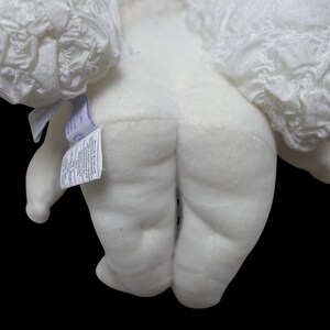 Puede incluir: Un juguete de peluche blanco con alas con volantes. El cuerpo del juguete es de un color blanco roto suave, con detalles cosidos. Una etiqueta est&aacute; adherida al juguete. El fondo es negro.