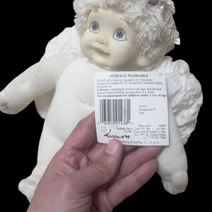 Puede incluir: Una mu&ntilde;eca Dreamsicles blanca con una cara sonriente, ojos azules y alas esponjosas. La mu&ntilde;eca tiene una etiqueta que dice "Surface Washable" y "Dreamsicles Doll". La mu&ntilde;eca es sostenida por una persona.