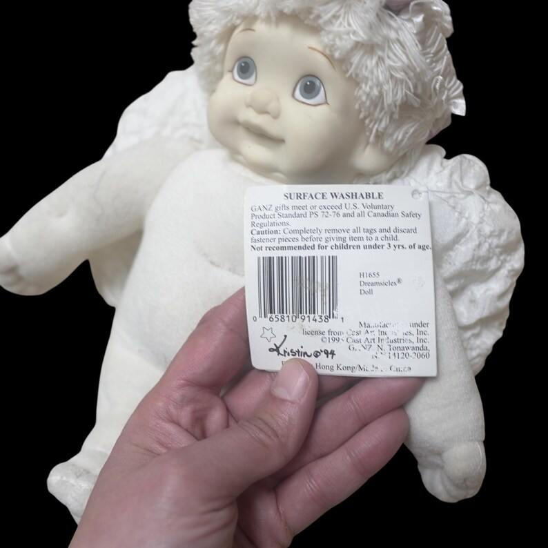 Puede incluir: Una mu&ntilde;eca Dreamsicles blanca con una cara sonriente, ojos azules y alas esponjosas. La mu&ntilde;eca tiene una etiqueta que dice "Surface Washable" y "Dreamsicles Doll". La mu&ntilde;eca es sostenida por una persona.