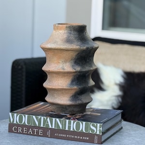 May include: A decorative, textured terracotta vase with a unique, ridged design. The vase has a natural, earthy color palette with dark gray accents. It sits atop two books with the title "MOUNTAIN HOUSE CREATE".