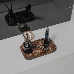 May include: A decorative tray with a marble pattern holds a black diffuser bottle with reeds, a jade roller, and a perfume bottle. A seashell is also on the tray. The items are arranged on a white surface.