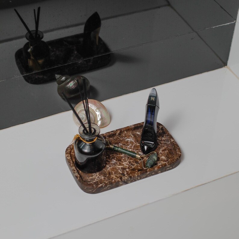 May include: A decorative tray with a marble pattern holds a black diffuser bottle with reeds, a jade roller, and a perfume bottle. A seashell is also on the tray. The items are arranged on a white surface.
