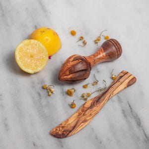 May include: A wooden citrus juicer and a butter knife with a natural wood grain pattern. A halved lemon and a whole lemon are next to the juicer. Dried chamomile flowers are scattered around the items.