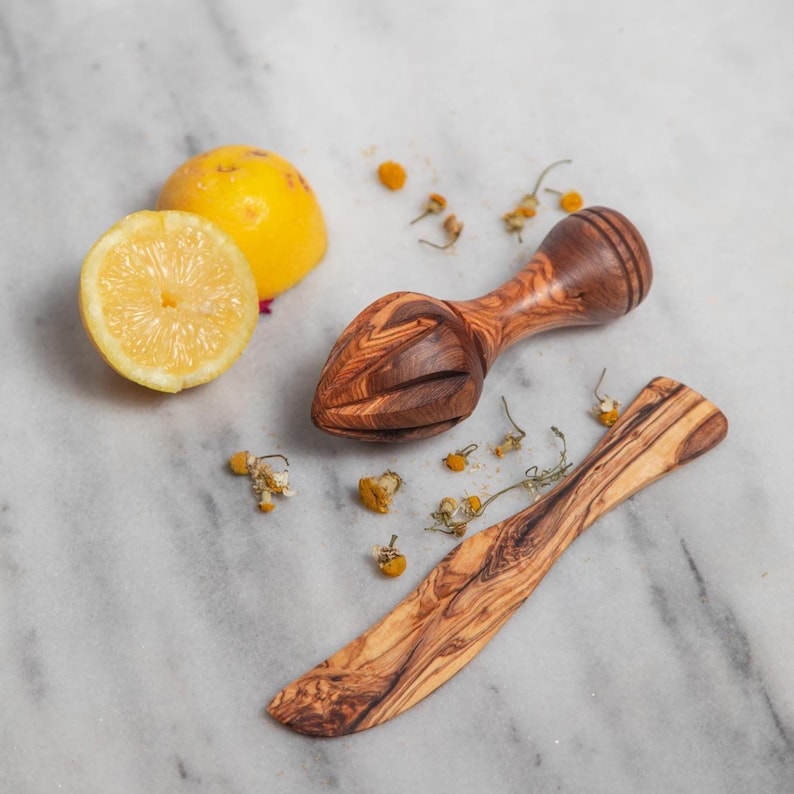 May include: A wooden citrus juicer and a butter knife with a natural wood grain pattern. A halved lemon and a whole lemon are next to the juicer. Dried chamomile flowers are scattered around the items.