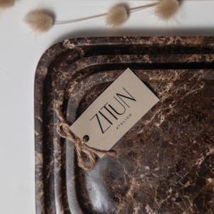 May include: Brown marble-patterned serving tray with rounded edges. A tag with the text "ZITUN ATELIER" is attached with twine. The tray has a dark brown and beige color scheme, with a marbled texture.