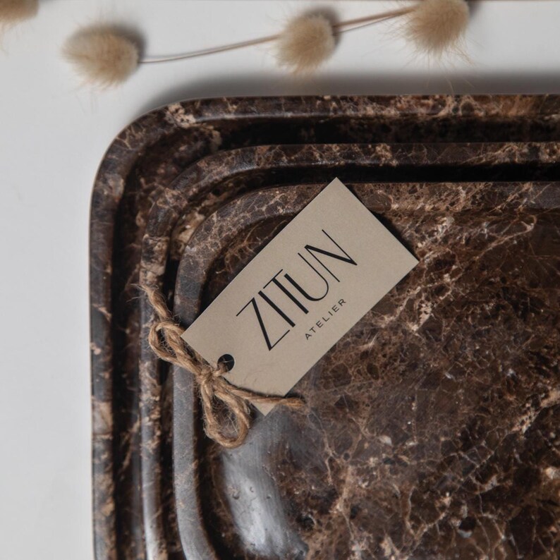 May include: Brown marble-patterned serving tray with rounded edges. A tag with the text "ZITUN ATELIER" is attached with twine. The tray has a dark brown and beige color scheme, with a marbled texture.