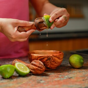 May include: A wooden citrus juicer is used to squeeze a lime, with juice dripping into a wooden bowl. Two additional juicers and two limes are on the counter. The juicers and bowls are made of olive wood.
