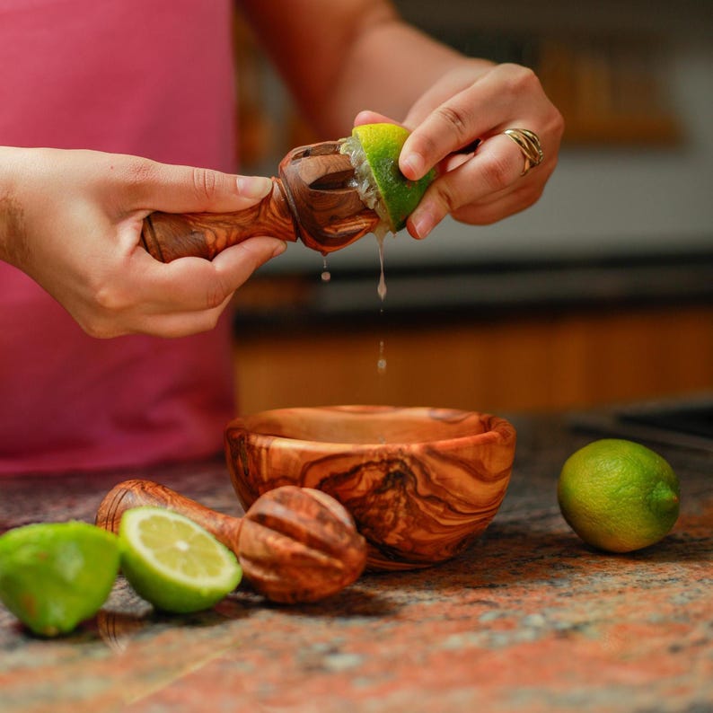 May include: A wooden citrus juicer is used to squeeze a lime, with juice dripping into a wooden bowl. Two additional juicers and two limes are on the counter. The juicers and bowls are made of olive wood.