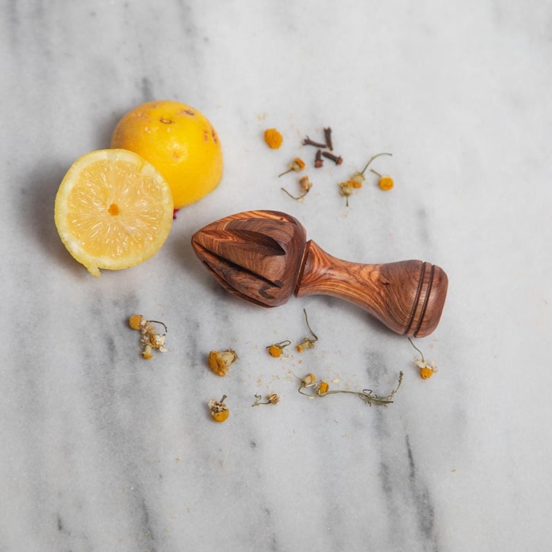May include: A wooden citrus juicer with a brown finish, next to a halved lemon and a whole lemon. Dried chamomile flowers and cloves are scattered around the juicer and lemons on a marble surface.