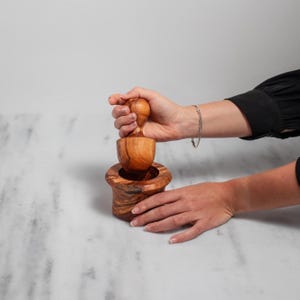 May include: A wooden mortar and pestle set. The mortar is a small, round bowl with a wide base and a textured exterior. The pestle is a rounded, mushroom-shaped piece. The set is made of a light brown wood and is being held by a person.