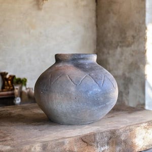 May include: A gray ceramic pot with a decorative pattern of triangles around the middle. The pot is sitting on a wooden surface.