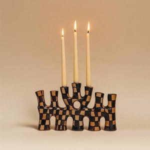 May include: A ceramic candelabra featuring a black and tan checkered pattern with an irregular, organic shape. The piece has three candle holders, each containing a lit, slender, off-white taper candle with a visible flame at the top.