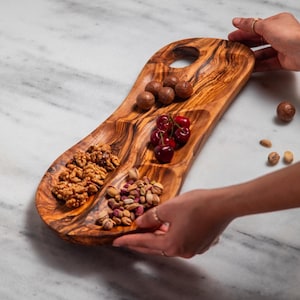 May include: A wooden serving tray with three compartments, filled with walnuts, pistachios, cherries, and macadamia nuts. The tray has a natural wood grain and a hole for hanging. The tray is being held by a person.